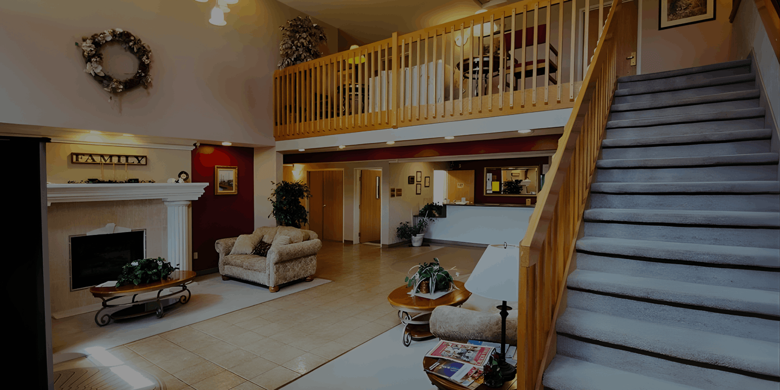 A large, open lobby or living area with a carpeted staircase leading to a mezzanine level, a seating area with a fireplace, and tan-colored furniture.