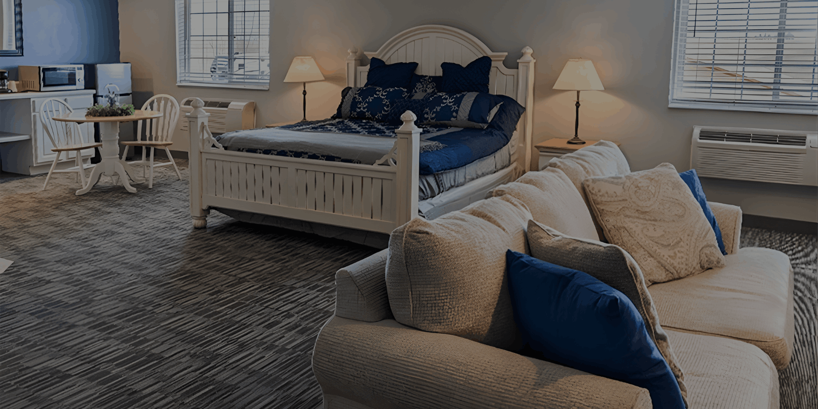 A spacious hotel suite featuring a white ornate bed with blue bedding, a tan sofa with blue pillows in the foreground, and a small dining area.