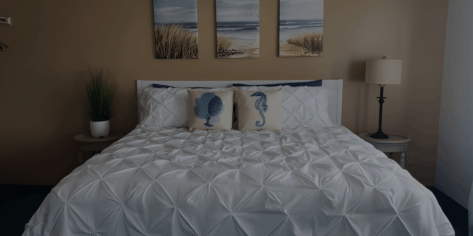 A bed with a white textured comforter and nautical-themed pillows positioned beneath three beach-themed landscape pictures on a tan wall.