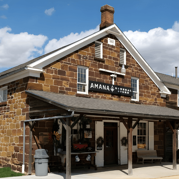 A historic two-story building made of brown fieldstone with a wooden front porch and a sign that reads "AMANA GENERAL STORE."