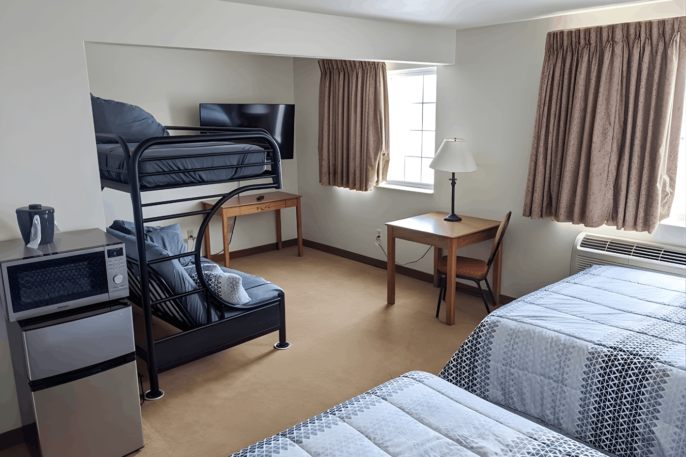 A multi-bed hotel room featuring two twin beds with patterned comforters, a black metal bunk bed, two wooden desks, and a kitchenette area with a mini-fridge and microwave.