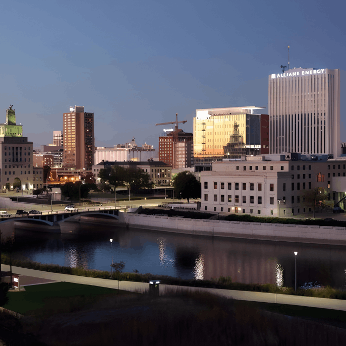 A city skyline at dusk with illuminated buildings reflecting in a calm river and a bridge in the background.