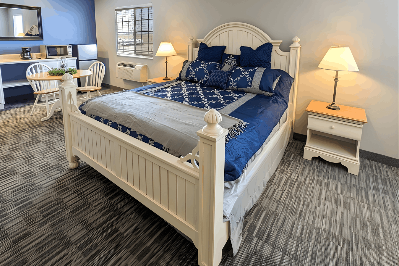 A hotel suite featuring a white wooden bed with blue-and-white patterned bedding, two matching nightstands with lamps, and a small dining area in the background.