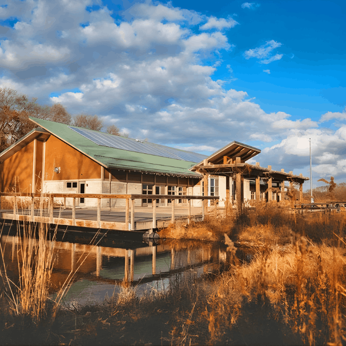 A modern wood-sided building with a slanted green roof next to a calm pond, featuring wooden walkways and reflections in the water.