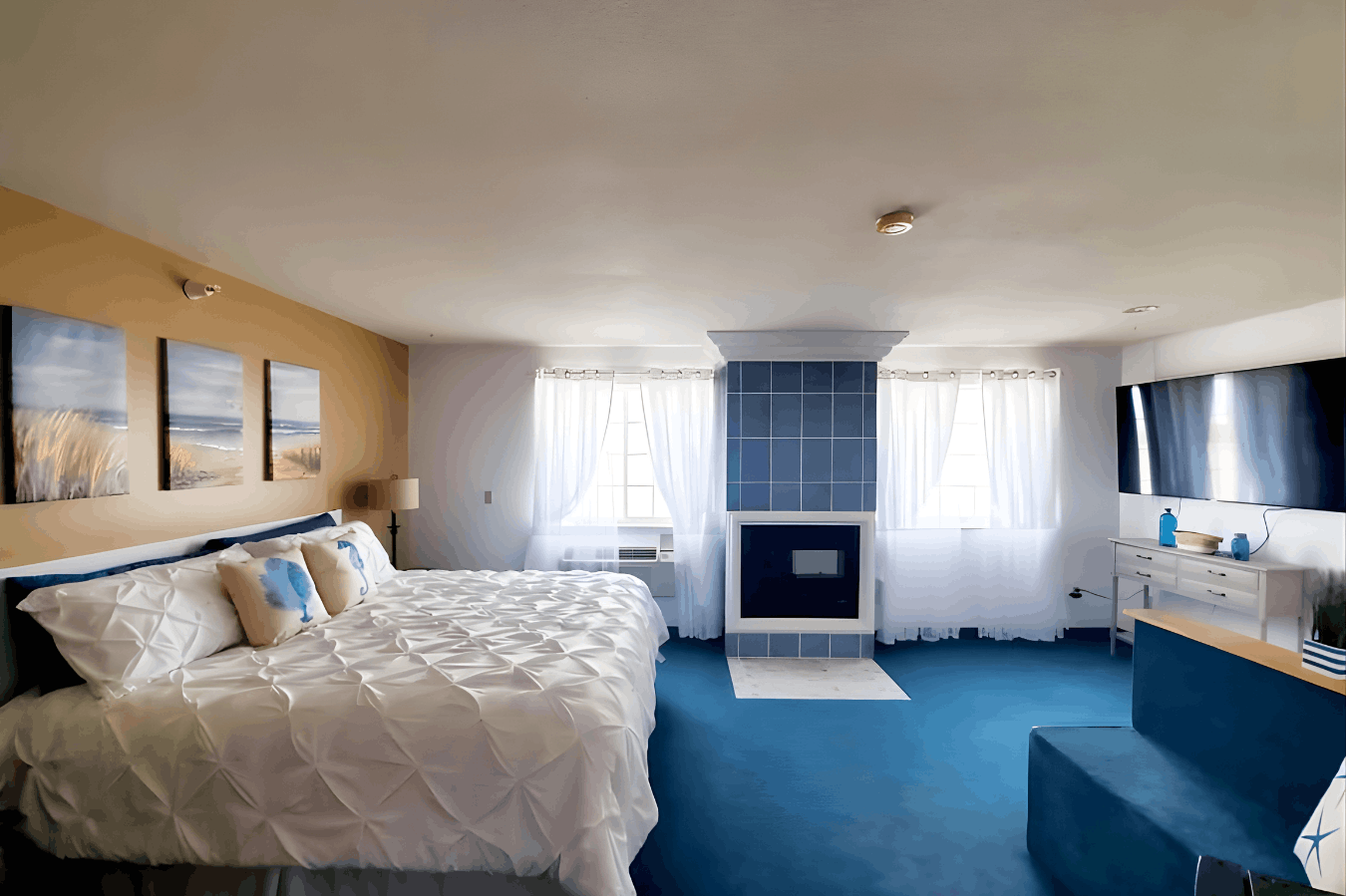 A spacious hotel room with coastal decor, blue carpeting, a white textured bed, and a blue-tiled fireplace column situated between two windows.
