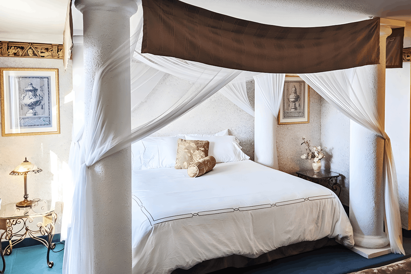A grand canopy bed featuring four large white classical pillars, draped with white sheer fabric and a brown top banner, set against white walls with framed art.