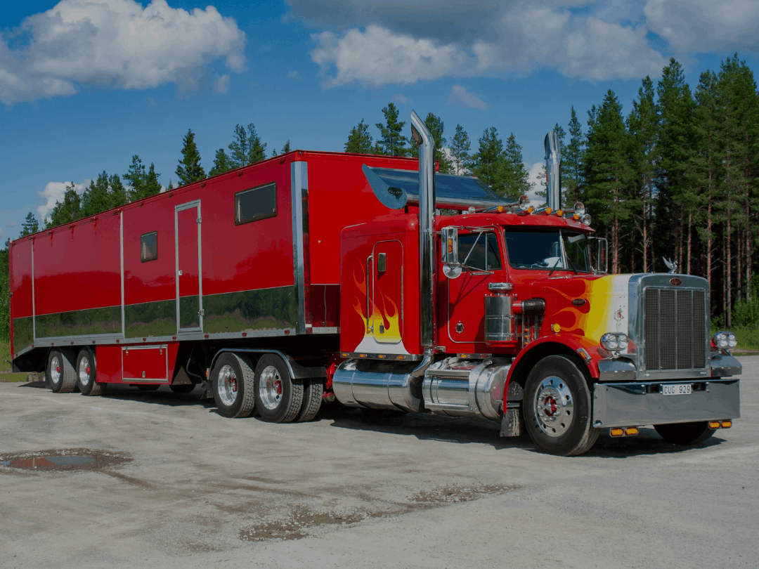 A vibrant red custom Peterbilt semi-truck with yellow and orange flame decals, pulling a large matching red trailer with chrome accents, parked on a gravel lot with a pine forest background.