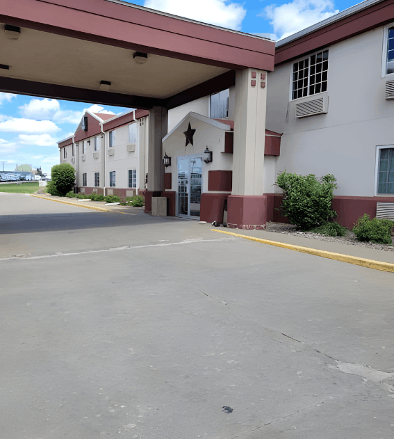 Exterior view of a two-story hotel featuring a large reddish-brown porte-cochère, a glass entrance with a star emblem, and light gray walls under a bright blue sky.