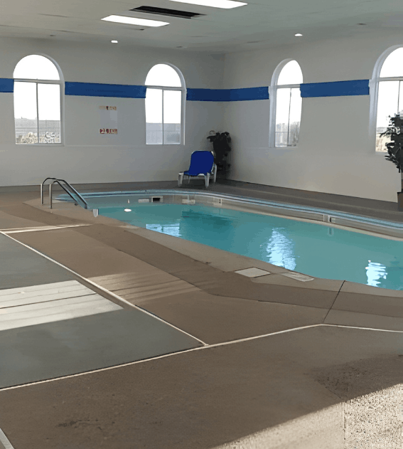An indoor swimming pool with clear blue water, surrounded by a tan deck, white walls with blue stripes, and large arched windows.